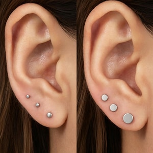 May include: A set of three silver-toned stud earrings arranged in a vertical line on an earlobe. The earrings are circular, flat, and polished, featuring a smooth metallic surface. The set displays three distinct sizes, graduating from small to large.