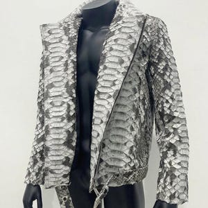 May include: A motorcycle-style jacket featuring a textured snakeskin pattern in shades of grey, white, and black. The jacket includes a diagonal front zip, a wide notched collar, long sleeves, and a matching belt with a metal buckle at the waist.