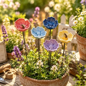 Può includere: Cinque picchetti da giardino a forma di fiore in ceramica rossa, azzurra, blu scuro, viola e gialla, montati su sottili aste di metallo nero. Due fiori presentano piccole statuette di api sui petali, il tutto in un vaso di terracotta.