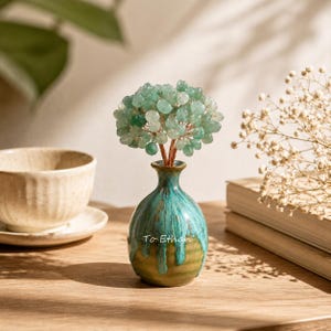 May include: A decorative tree sculpture with light green gemstone leaves on copper wire branches, set in a small ceramic vase with a teal and tan drip glaze finish. The vase features the white text "To Ethan" printed on the front surface.