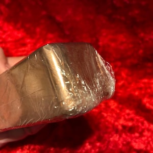 May include: A rectangular copper-colored metal bar with rounded edges, encased in a clear, crinkled plastic film wrapping. The surface of the metal has a smooth, metallic finish with a warm, reddish-brown hue visible through the transparent plastic.