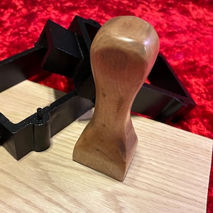 May include: A smooth, light brown wooden handle with a rounded top and a flared base, positioned next to a black, rectangular, hollow frame component. The wooden handle features a natural grain pattern and a polished finish, standing upright.