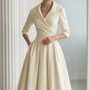 May include: A cream-colored midi dress featuring a wide shawl collar, a wrap-style bodice with subtle pleating, three-quarter length sleeves, and a full, pleated skirt. The dress includes a defined waistline and side pockets in a solid fabric.