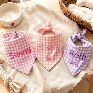 May include: Three triangular pet bandanas featuring a gingham check pattern. The left bandana is pink with the embroidered name Sunny. The center bandana is light pink with the name Heath and a paw print. The right bandana is purple with the name Roth and a bone.