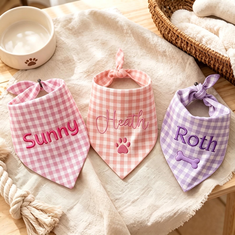 May include: Three triangular pet bandanas featuring a gingham check pattern. The left bandana is pink with the embroidered name Sunny. The center bandana is light pink with the name Heath and a paw print. The right bandana is purple with the name Roth and a bone.