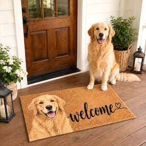 Peut inclure: Un paillasson rectangulaire en fibre de coco beige avec une image imprimée d'une tête de golden retriever sur la gauche et le mot welcome en écriture cursive noire avec une petite icône de cœur sur la droite, posé sur un sol en bois.