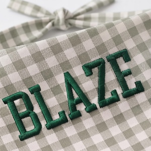 May include: A sage green and white gingham fabric accessory featuring the word BLAZE embroidered in dark green capital letters. The item includes a tied fabric strap at the top, showcasing a classic checkered pattern and textured stitching.