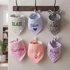 May include: Six triangular pet bandanas in various gingham patterns, each featuring embroidered names and icons. The bandanas are colored grey, light blue, black, pink, peach, and purple, and are tied to a dark metal wall-mounted hook rack.