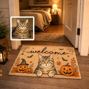 Op de afbeelding: Een rechthoekige, lichtbruine deurmat met een centrale illustratie van een bruine cyperse kat met groene ogen, geflankeerd door twee oranje pompoenen. De mat bevat het woord welcome in zwarte letters, spinnenwebben, vleermuizen en herfstbladeren.