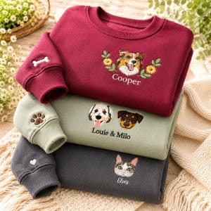 Könnte beinhalten: Ein Stapel aus drei Rundhals-Sweatshirts in Kastanienbraun, Salbeigrün und Anthrazit. Jedes zeigt gestickte Haustierporträts und Namen: Cooper mit Blumen, Louie und Milo sowie Elvis. Die Ärmel zeigen ein Knochen-, Pfoten- und Herzsymbol.