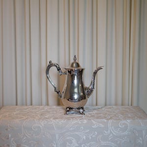 May include: A polished silver-tone metal coffee pot featuring an ornate, curved handle, a decorative spout, and a matching lid with a small finial. The vessel stands on four detailed, scrolled feet and has a smooth, reflective surface throughout.