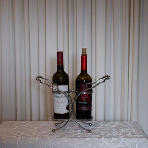 May include: A silver-toned metal wine rack holding two glass bottles. The left bottle features a white label with the text Stella Rosa, while the right bottle has a dark label with the text Apothic Red. The rack has a curved, open-frame design.
