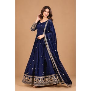 Puede incluir: Un vestido anarkali azul marino largo hasta el suelo con mangas largas, cuello redondo y corpiño ajustado. La tela está adornada con pequeños motivos florales dorados y un borde ancho y complejo bordado en oro a lo largo del dobladillo y los puños.
