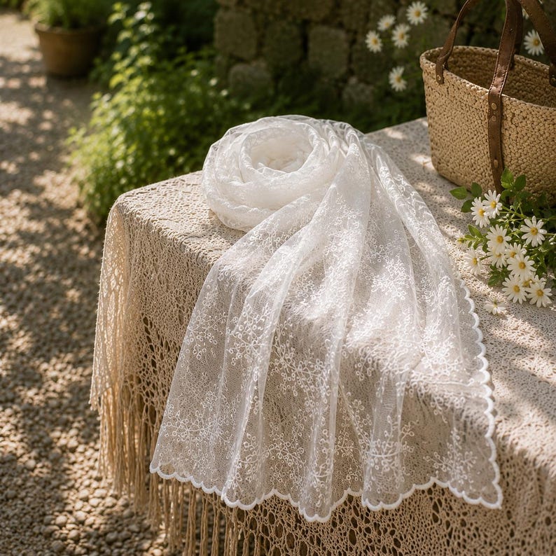 May include: A white rectangular scarf featuring an intricate floral lace pattern with a scalloped edge. The lightweight fabric is semi-sheer and displays a delicate embroidered design throughout the entire length of the accessory.