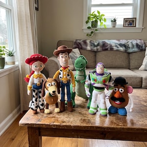 Könnte beinhalten: Eine Sammlung von fünf gehäkelten Puppen, die Toy Story-Figuren darstellen, darunter eine Cowgirl mit rotem Hut, ein brauner Dackel, ein Sheriff mit braunem Hut, ein grüner Dinosaurier, ein Weltraum-Ranger und eine kartoffelförmige Figur mit schwarzem Hut.
