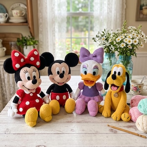 May include: Four handmade crochet dolls featuring classic cartoon characters: a mouse in a red polka-dot dress with a matching bow, a mouse in red shorts, a duck in a purple dress with a matching bow, and a yellow dog with black ears.