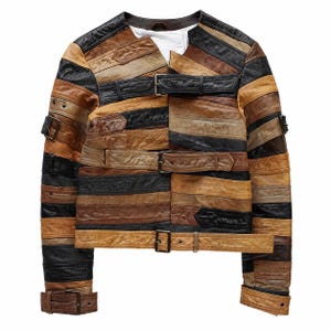 Puede incluir: Una chaqueta confeccionada con tiras horizontales de cuero negro, tostado y marrón. La prenda presenta un escote sin cuello, mangas largas con puños de hebilla y tres correas horizontales con hebilla en el cierre frontal. Se observa un forro blanco.