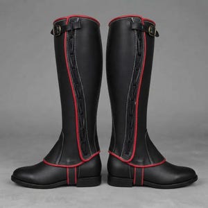 May include: A pair of black knee-high boots featuring red piping along the edges, white contrast stitching, and a vertical lace-up design on the outer side. Each boot includes a black strap with a metal buckle at the top for a secure fit.