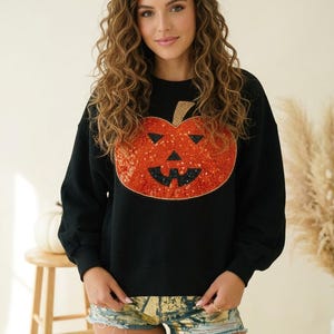 May include: A black long-sleeve sweatshirt featuring a large, central pumpkin graphic made of orange and black sequins. The pumpkin has a jack-o'-lantern face with triangular eyes and a jagged mouth. The sweatshirt has a crew neckline and ribbed cuffs.
