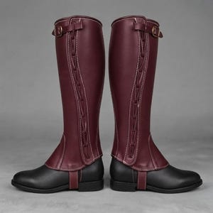 May include: A pair of knee-high gaiters featuring a deep burgundy upper section with decorative side lacing and a buckle strap at the top, paired with a solid black lower foot section that includes a vertical strap across the instep area.