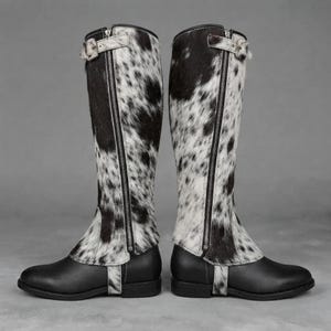 May include: A pair of knee-high boots featuring a black and white cowhide pattern on the shaft with a vertical silver zipper closure. The lower portion of the boots is constructed from smooth black leather with a matching cowhide strap detail.
