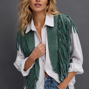 Op de afbeelding: Een donkergroen suède vest met lange franjes langs de voorpanden en schouders. Het vest wordt gedragen over een wit overhemd met knoopsluiting en gecombineerd met een blauwe spijkerbroek, wat zorgt voor een gelaagde look met textuur.