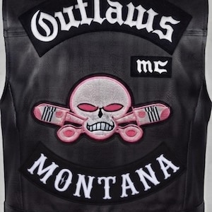 May include: A black leather vest featuring embroidered patches on the back. The top patch reads Outlaws in white gothic lettering, with a smaller MC patch below. A central skull emblem with pink accents and crossed pistons sits above a Montana patch.