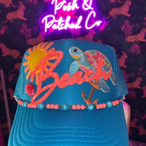 Peut inclure: Une casquette trucker bleu sarcelle ornée d'un soleil jaune brodé, d'une tortue de mer colorée, d'un petit crabe rouge et d'une ancre marron. Le mot Beach est écrit en grande écriture orange pailletée. Une bordure de perles corail et bleues orne la visière.