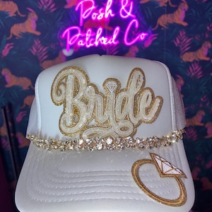 May include: A white trucker hat featuring the word Bride in gold sequined script lettering across the front panel. A row of clear rhinestones sits above the brim, and a gold sequined diamond ring patch is positioned on the right side of the brim.