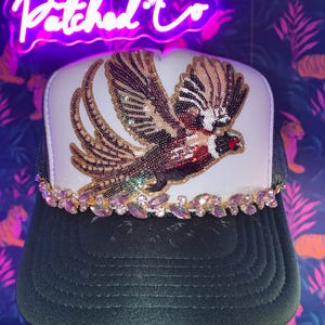 May include: A white and black trucker hat featuring a large, sequined pheasant patch on the front panel. The base of the front panel is adorned with a decorative trim of iridescent, light purple, marquise-cut rhinestones set in a gold-tone metal.