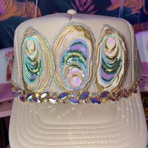 May include: A beige trucker hat featuring three embroidered oyster shell patches on the front panel. Each shell is embellished with iridescent sequins in shades of green, pink, and gold. A row of faceted, purple-toned crystals lines the brim base.