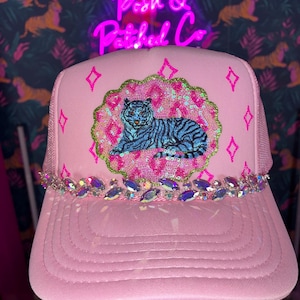 May include: A light pink trucker hat featuring a central circular patch with a blue tiger graphic on a pink sequined background. The hat is adorned with a row of iridescent rhinestones across the brim and small pink star-shaped accents on the front panel.