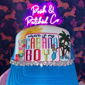 Peut inclure: Une casquette de camionneur bleu vif avec un panneau avant blanc et un écusson brodé portant l'inscription where is my CABANA BOY en lettres colorées. Le motif inclut un soleil jaune, des palmiers et des cocktails, orné de strass transparents.