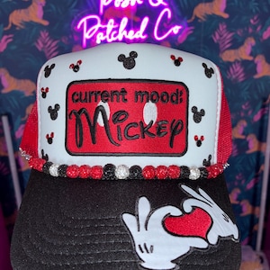 May include: A white and black trucker hat featuring a red rectangular patch with the text current mood: Mickey. The hat is decorated with small black Mickey Mouse head icons, a row of red, black, and white beads, and a graphic of white gloved hands forming a heart shape over a red heart on the black brim.