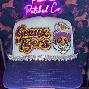 May include: A navy blue and white trucker hat featuring a sequined patch that reads Geaux Tigers in purple and gold. Next to the text is a patch of a tiger wearing a purple cap and star-shaped glasses. A row of clear rhinestones lines the brim.