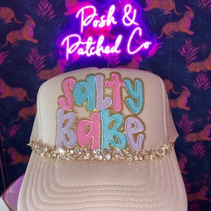 Peut inclure: Une casquette de type trucker couleur crème avec l'inscription "Salty Babe" en lettres bulles scintillantes roses et bleues sur un fond doré pailleté. Une rangée de strass transparents étincelants est fixée à la base du panneau avant.