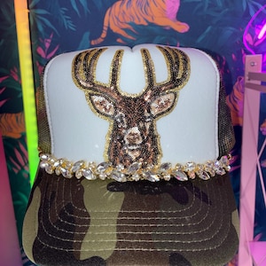 Puede incluir: Una gorra tipo trucker con panel frontal blanco y un diseño de cabeza de ciervo con lentejuelas en tonos marrón, negro y dorado. Una hilera de cristales transparentes facetados bordea la base del panel frontal y la visera tiene un patrón de camuflaje verde y marrón.
