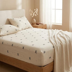Puede incluir: Un juego de sábanas y fundas de almohada de color crema con un patrón repetitivo de pequeños lazos y puntos negros. La ropa de cama está dispuesta sobre una cama con cabecero claro, acompañada de una manta de color crema lisa sobre el lateral.