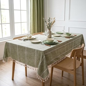 May include: A rectangular sage green tablecloth with a white vertical striped pattern and decorative fringe along the edges. The tablecloth is draped over a wooden dining table set with white plates, bowls, and clear glassware for four people.