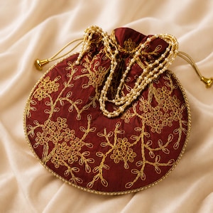 May include: A round, deep red drawstring pouch featuring intricate gold floral embroidery across the front. The bag is accented with a delicate beaded pearl handle and gold-toned drawstring cord ends, creating a structured yet soft appearance.