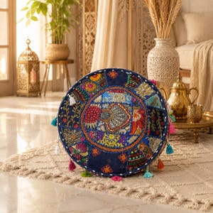 May include: A round floor cushion featuring a dark blue fabric base with a patchwork design of embroidered floral and geometric patterns in vibrant colors, accented with small mirrors, metallic thread, and colorful tassels along the edge.