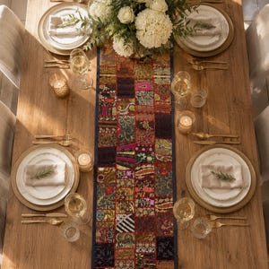 May include: A rectangular table runner featuring a patchwork design of various fabrics in shades of dark red, gold, black, and pink with intricate embroidery and patterns, centred on a wooden dining table with white plates and gold cutlery.