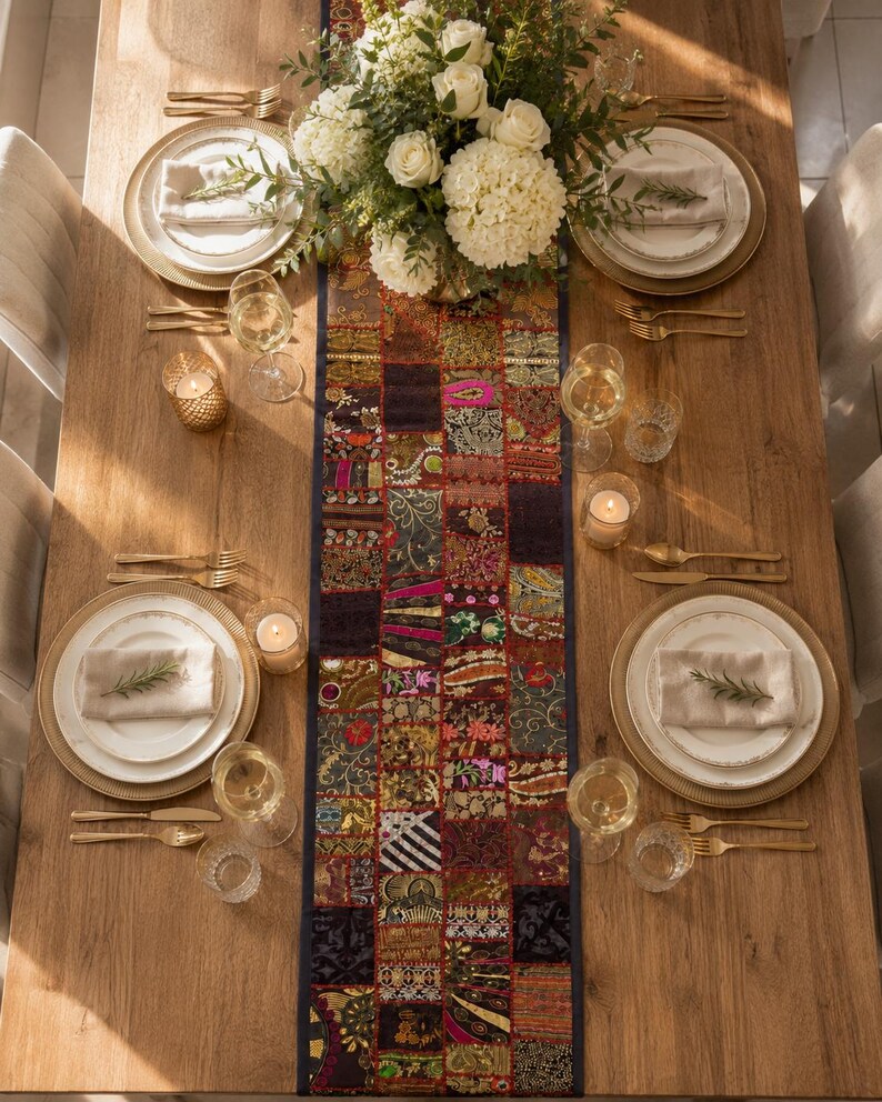 May include: A rectangular table runner featuring a patchwork design of various fabrics in shades of dark red, gold, black, and pink with intricate embroidery and patterns, centred on a wooden dining table with white plates and gold cutlery.