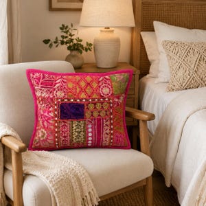 May include: A square throw pillow featuring a vibrant patchwork design in shades of hot pink, magenta, and gold. The surface is decorated with intricate embroidery, metallic thread accents, small circular mirrors, and various floral and geometric patterns.