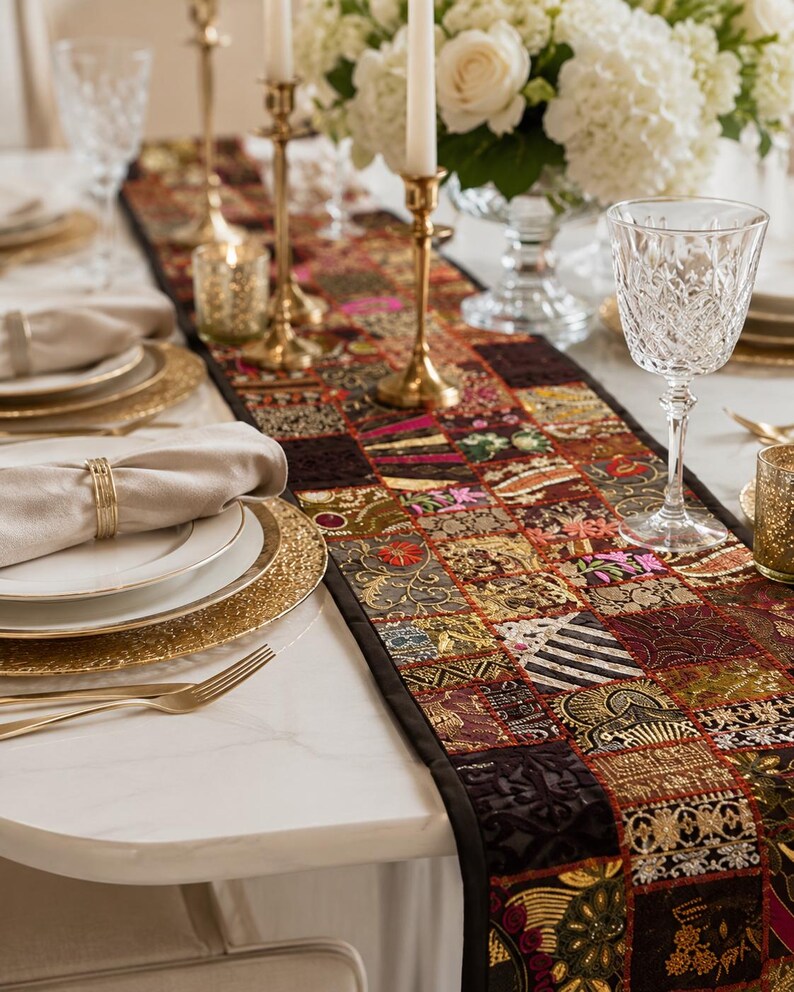 May include: A rectangular table runner featuring a patchwork design of various dark-toned fabric squares with intricate gold, red, and green embroidery, metallic thread accents, and floral patterns, finished with a solid black fabric border.