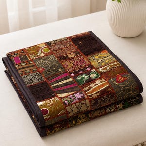 May include: A folded patchwork quilt featuring a variety of square fabric panels in dark brown, gold, green, and magenta tones. The quilt displays intricate embroidery, floral patterns, and textured stitching, finished with a solid dark brown border.