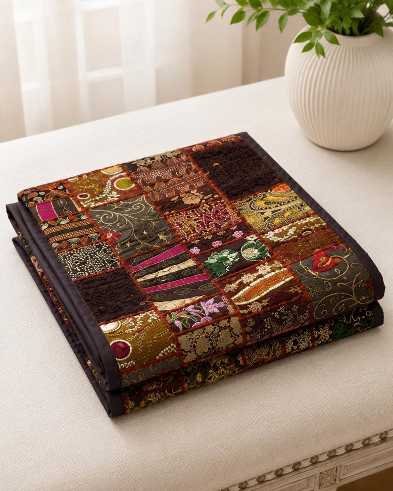 May include: A folded patchwork quilt featuring a variety of square fabric panels in dark brown, gold, green, and magenta tones. The quilt displays intricate embroidery, floral patterns, and textured stitching, finished with a solid dark brown border.