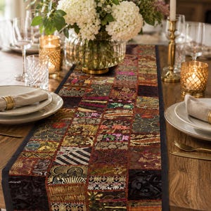 May include: A rectangular table runner featuring a patchwork design of various fabric squares in shades of black, gold, red, and green. The runner displays intricate embroidery, metallic threads, and diverse patterns, finished with a solid black border.