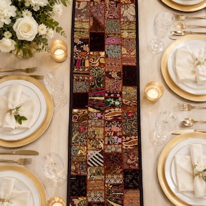 May include: A rectangular table runner featuring a patchwork design of various dark-toned fabric squares with intricate gold, red, and black embroidery, sequins, and metallic thread accents, centred on a light-coloured table with place settings.