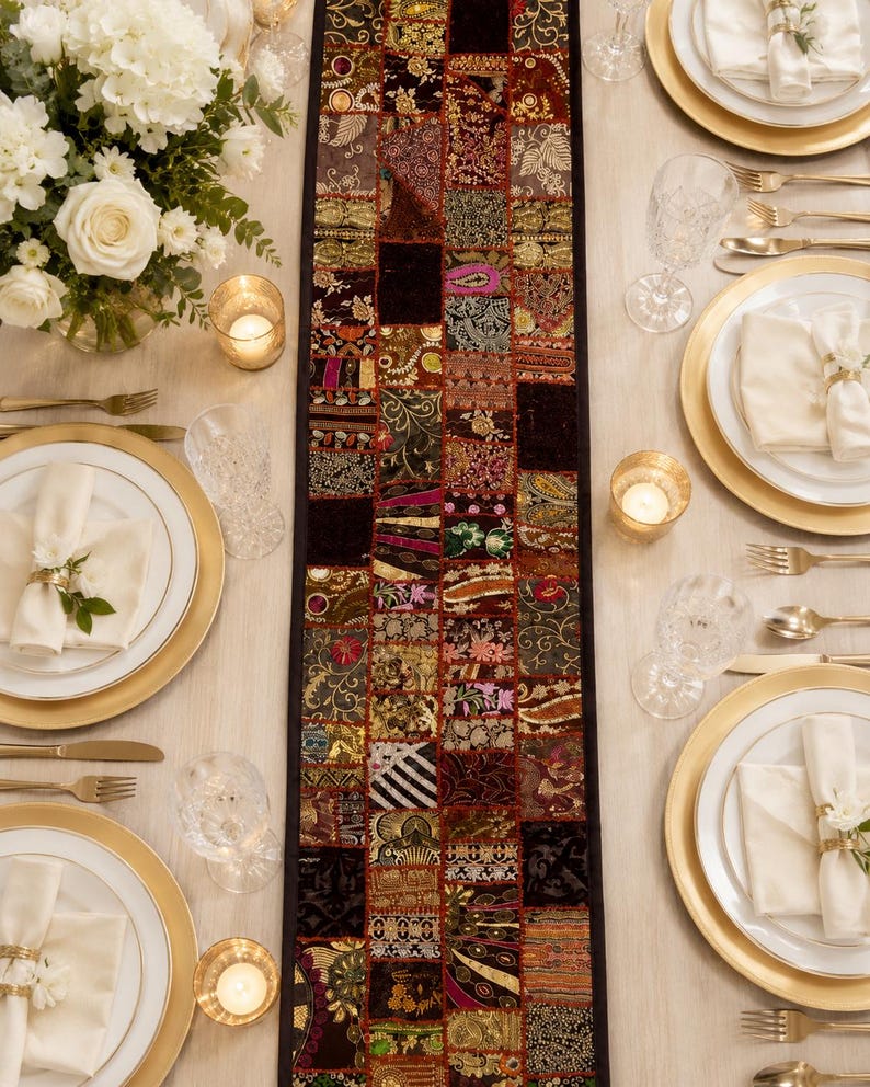 May include: A rectangular table runner featuring a patchwork design of various dark-toned fabric squares with intricate gold, red, and black embroidery, sequins, and metallic thread accents, centred on a light-coloured table with place settings.